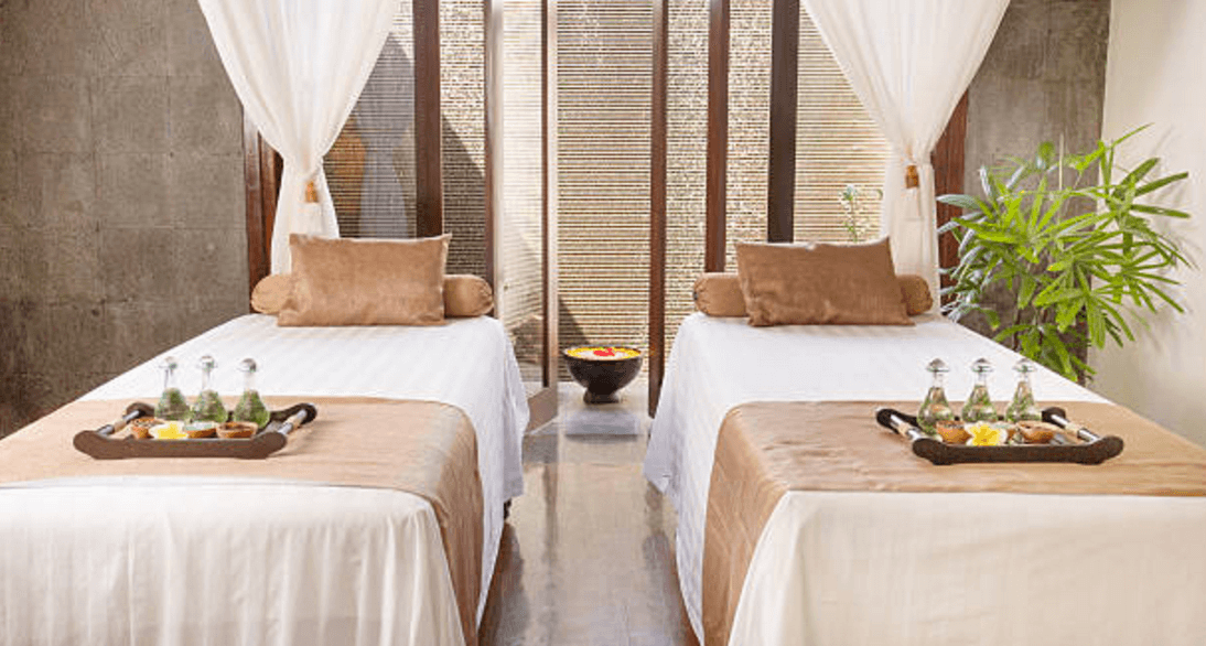 Two massage tables with beige linens and brown pillows, each with a tray holding bottles and snacks, in a bright spa room with tall windows and a green plant.