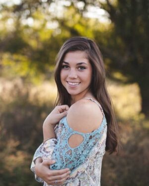 A young woman with long brown hair stands outdoors in a patterned top with shoulder cut-outs, smiling at the camera with trees and grass in the background.
