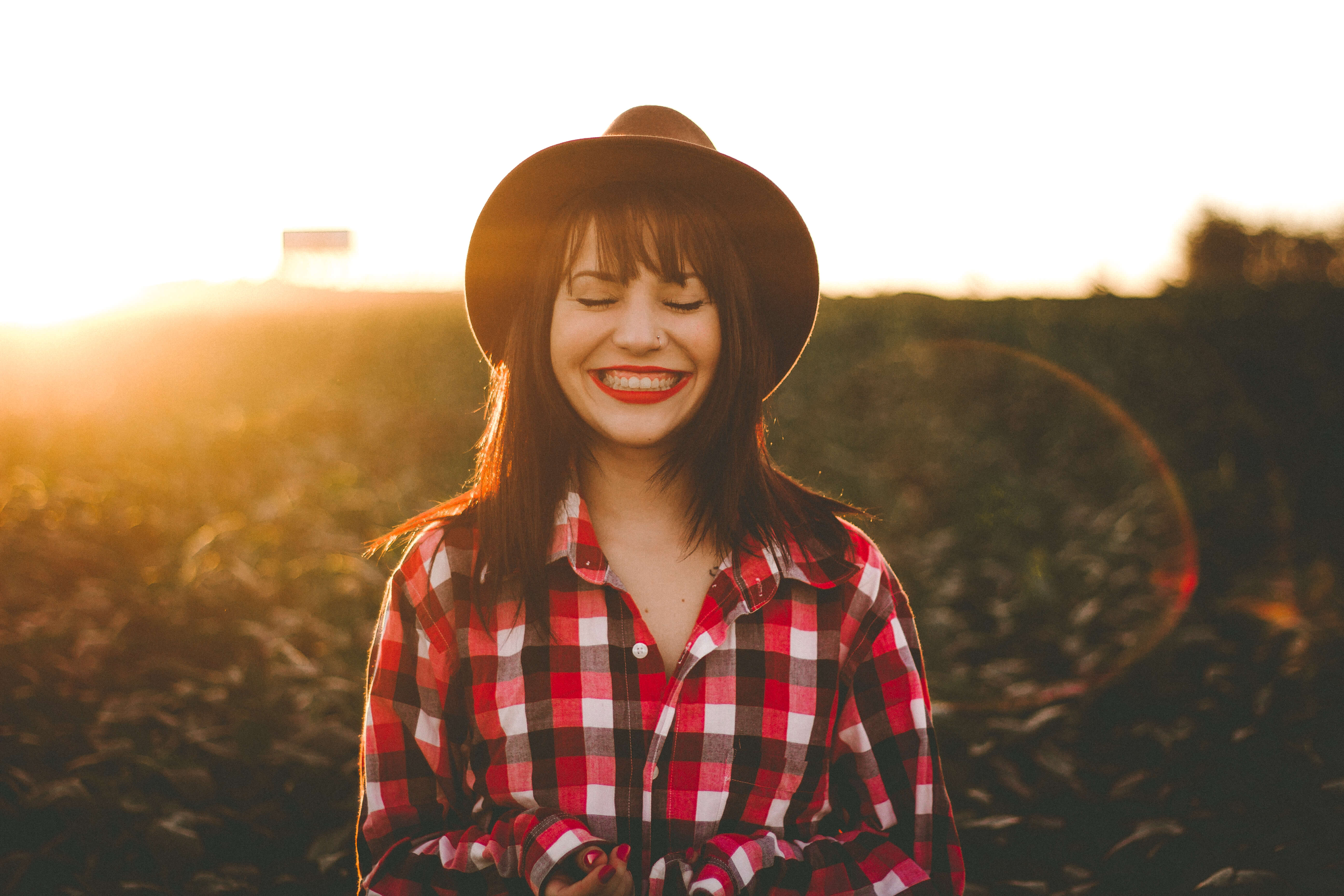 A person wearing a black hat and red plaid shirt stands outdoors in a field at sunset, smiling with eyes closed.